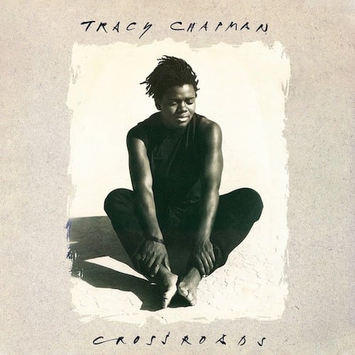 A slightly sepia-toned photo of Tracy Chapman, a young Black woman with short dreadlocks, sittng on sand. She holds her ankles. She wears a sleeveless black shirt, black pants, and is barefoot. Her eyes are closed, as if thinking hard or maybe just really really experiencing the sun reflecting on her.