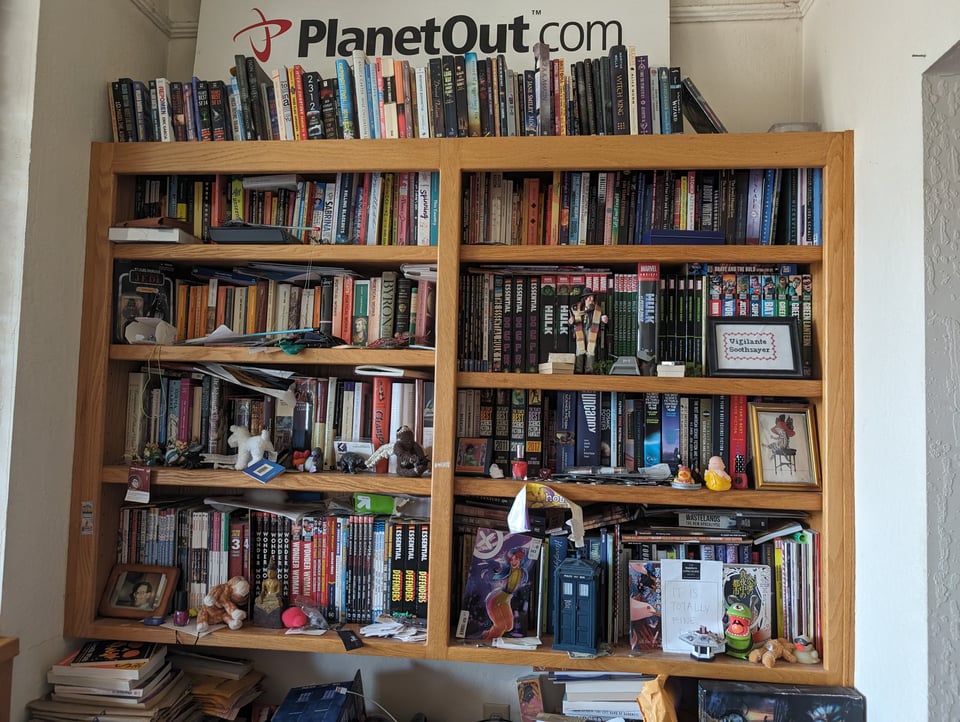 A bookshelf full of graphic novels, books, Doctor Who toys and other random clutter