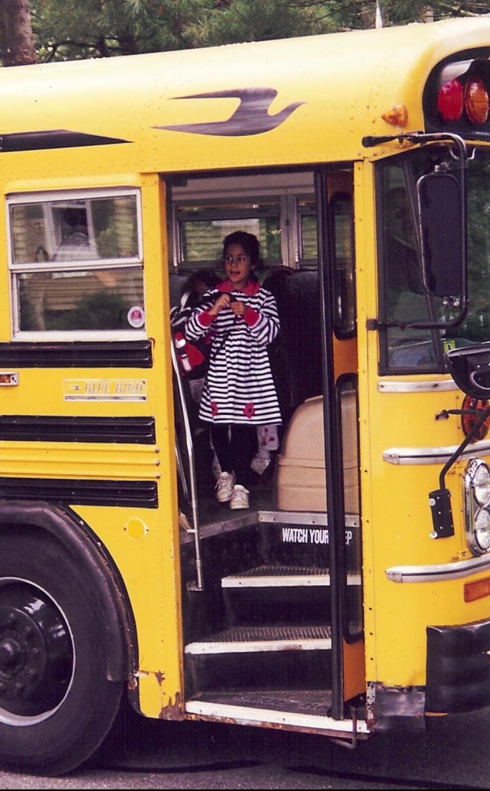 a 7-year-old Shivani stands at the top of the steps, about to descend from a yellow school bus. she's wearing a black and white striped dress with red flowers and holding a black, red, and white backpack.