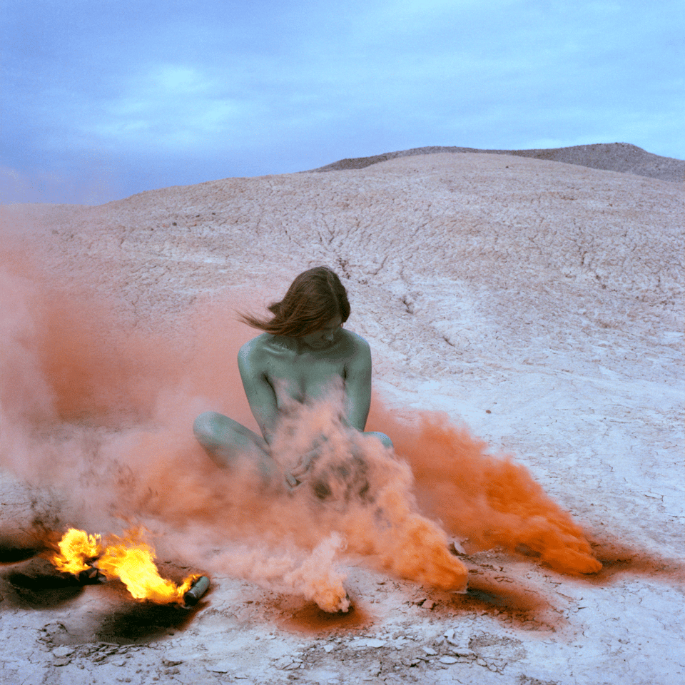 a woman covered in green body paint, sitting in the desert, surrounded by small fires and plumes of red smoke