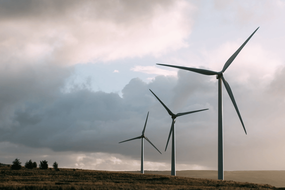 A photo of three windmills in a wide, empty field, against a sky, mostly covered in low, flying gray clouds with a bit of blue sunny sky in the center.