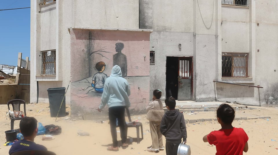 A photo from behind of a group of little kids gathered around the mural of the child looking at his shadow, without any text painted yet. There are bags and puts of ink on the ground.