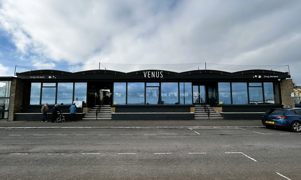 What looks like a single storey building facing a road. It has five concrete wave sections on the roof and large clear windows. Under the second and fourth concrete spans, steps rise up from the pavement to the entrance doors.