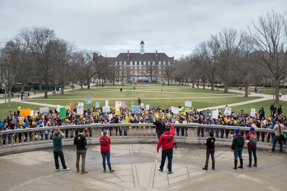Foreground: A crowd of striking graduate workers stands in front of the Foellinger Auditorium at the University of Illinois Urbana-Champaign. They are carrying protest signs and banners that describe their demands.

Background: The Main Quad of the University, a broad green space with criss-crossing sidewalks, and the Illini Union, a four-story colonial-style building.
