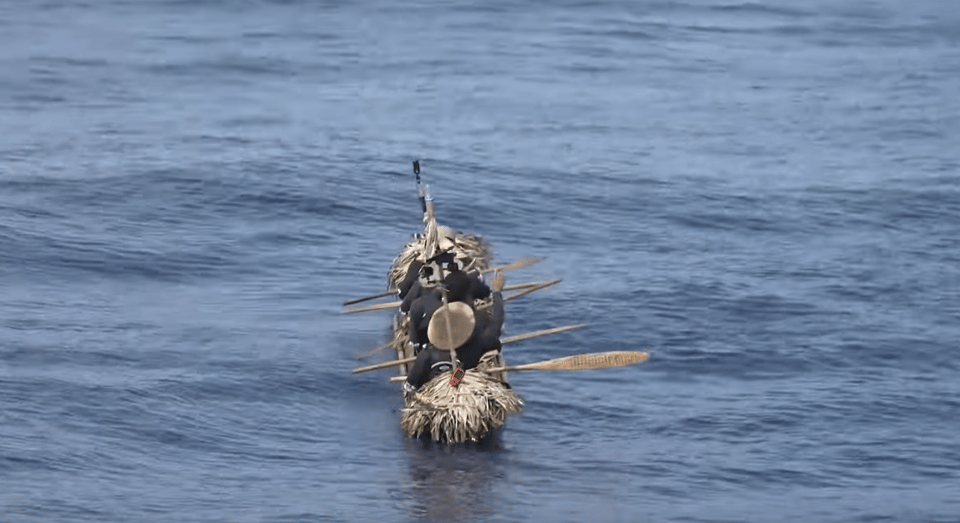 A photo of a narrow handmade canoe floating on open ocean water. It is pointed away from us, towards the horizon. It looks like it's been built from bundles of reeds or similar plant material. There are several people in the boat, all wearing wide-brimmed straw hats and holding wooden oars horizontally. They're not rowing right now. It looks very rugged