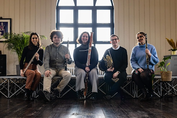 Portrait of musicians holding their wind instruments, seated in front of a church window.