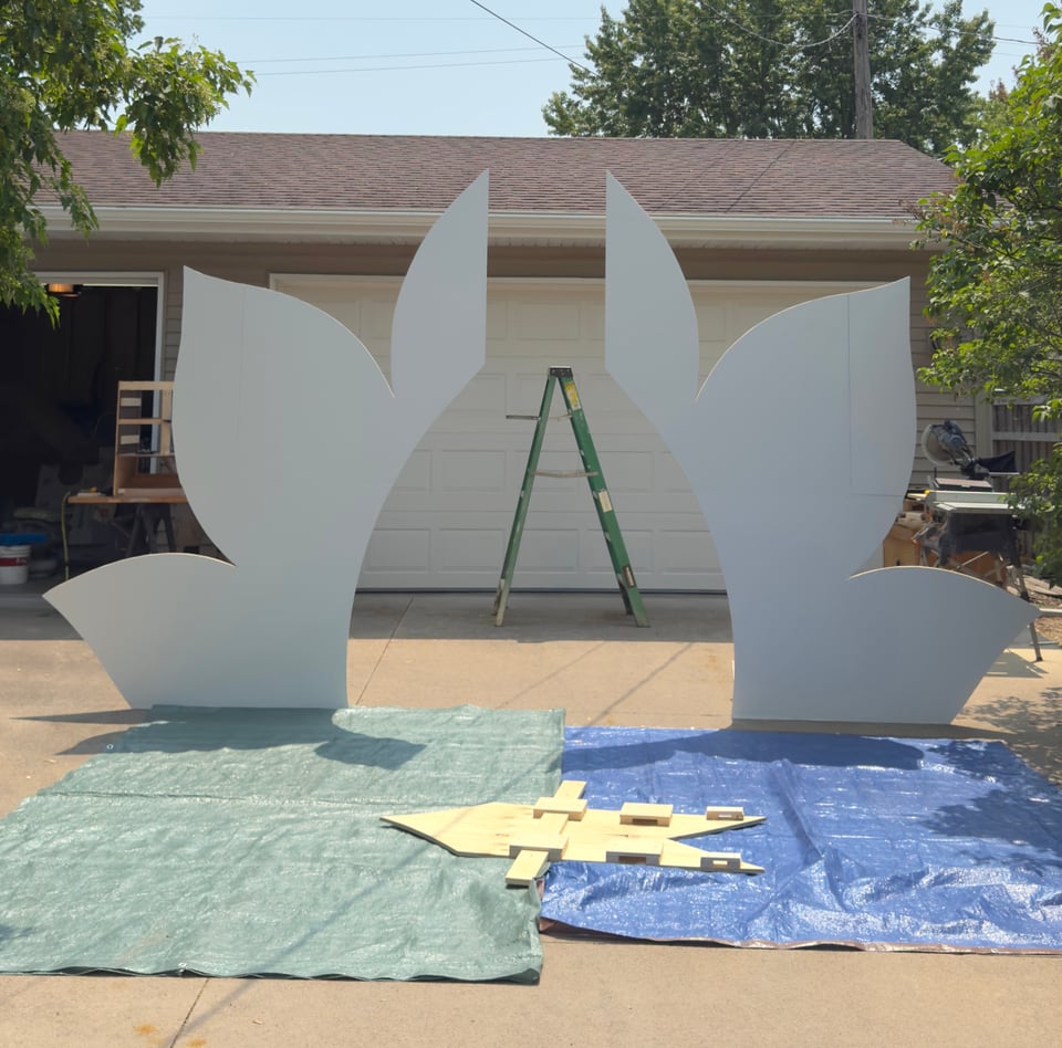 White arch display standing on tarps on a driveway with the center piece laying in front of it on the ground.