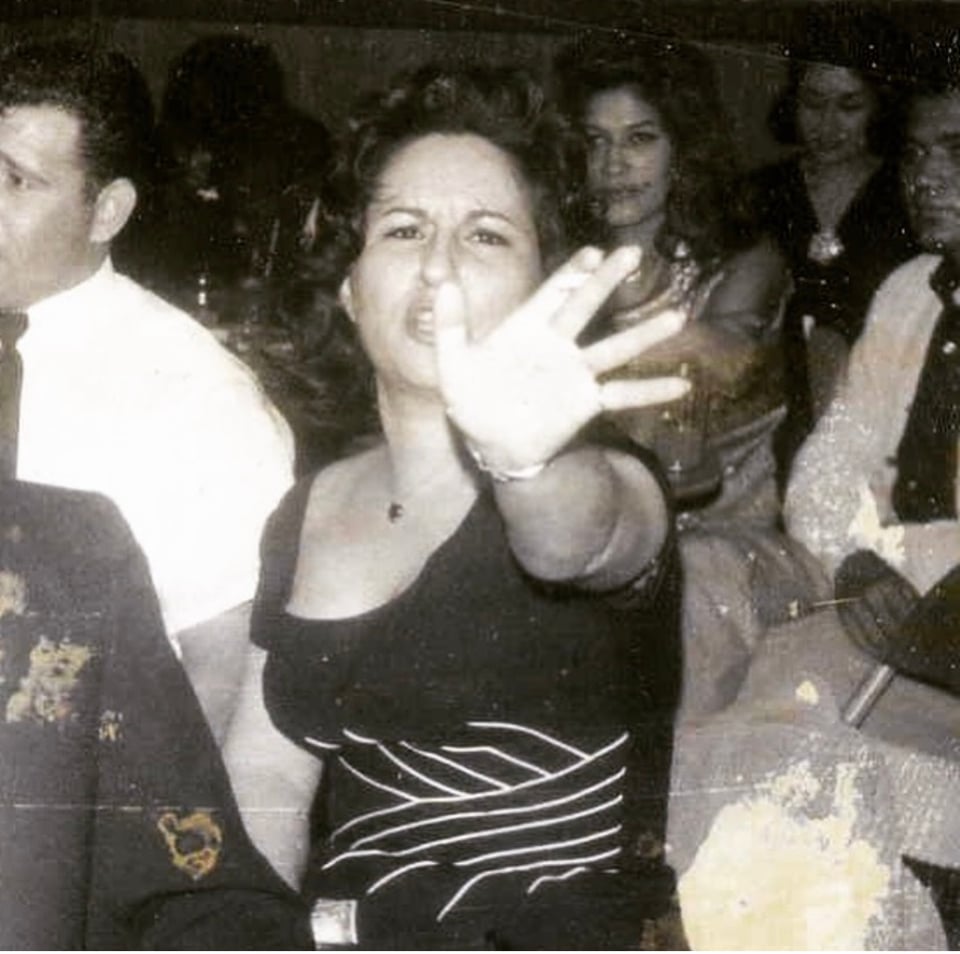 An old black and white photo of a woman who appears to be saying STOP to the photographer, her arm outstretched. She's wearing a black dress with white lined accents. She is in a nightclub at a table and you can see people sitting behind her. Parts of the photo are damaged with white spots.