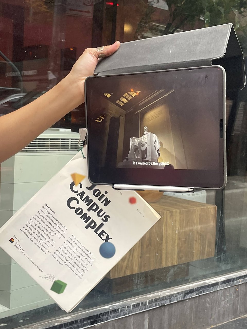 A hand holding an iPad with a still of the Jefferson Memorial and the subtitles “It's owned by the public.” Hanging underneath the iPad is a pamphlet that says Join Campus Complex.