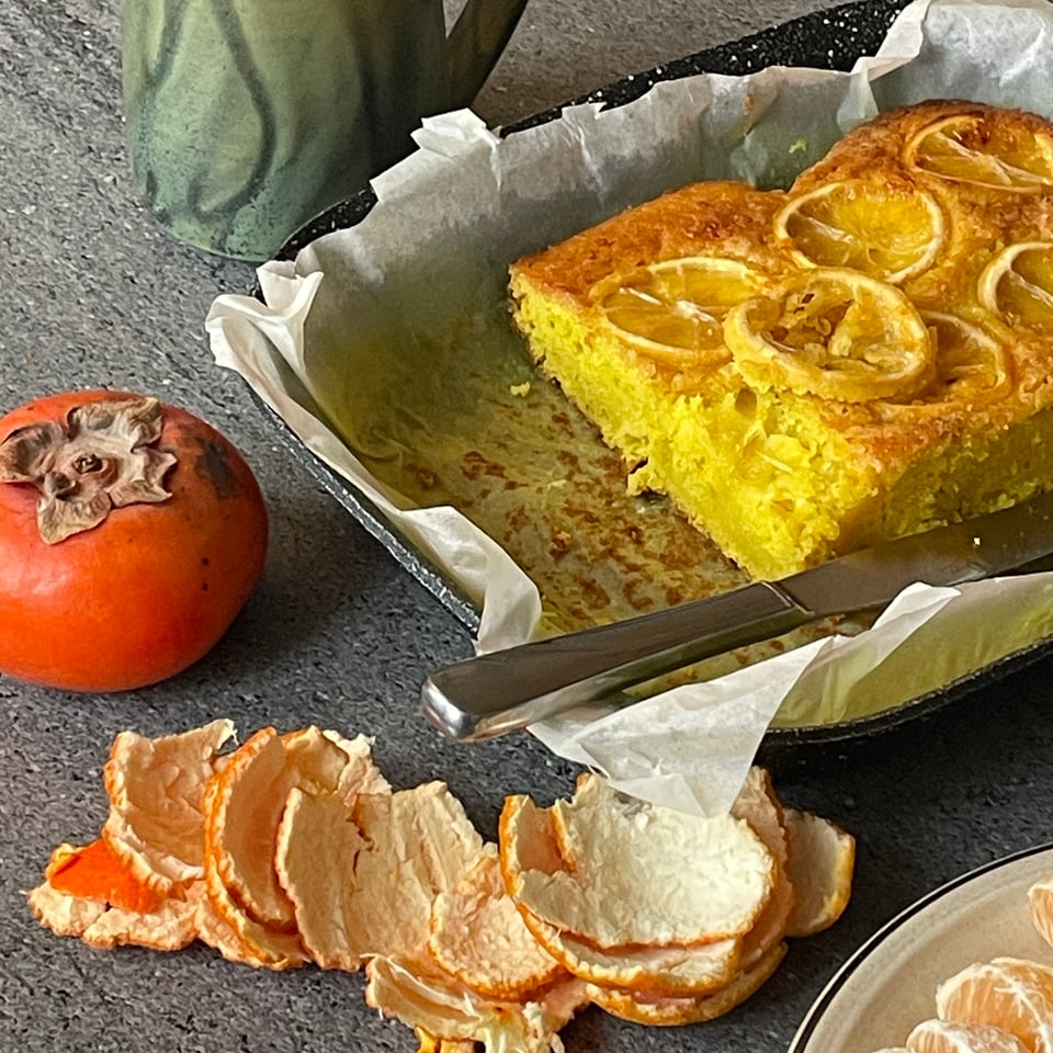 image of a persimmon, a clementine and its peels, a green handled mug, and a lemon cake with lemon slices atop it.