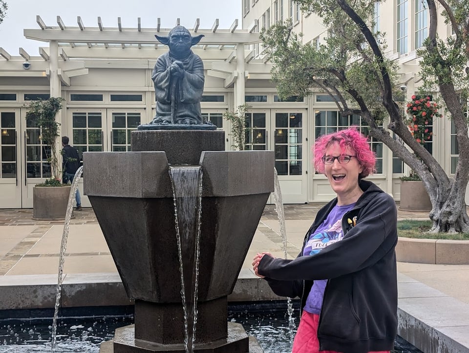 A statue of Yoda from Star Wars looking wise and stern, over a fountain that vents into a pool. Behind Yoda is a nice building with huge windows, and a tree. In front of Yoda, a trans woman with pink hair wearing a black hoodie, purple T-shirt and pink pants pretends to swing a light saber.