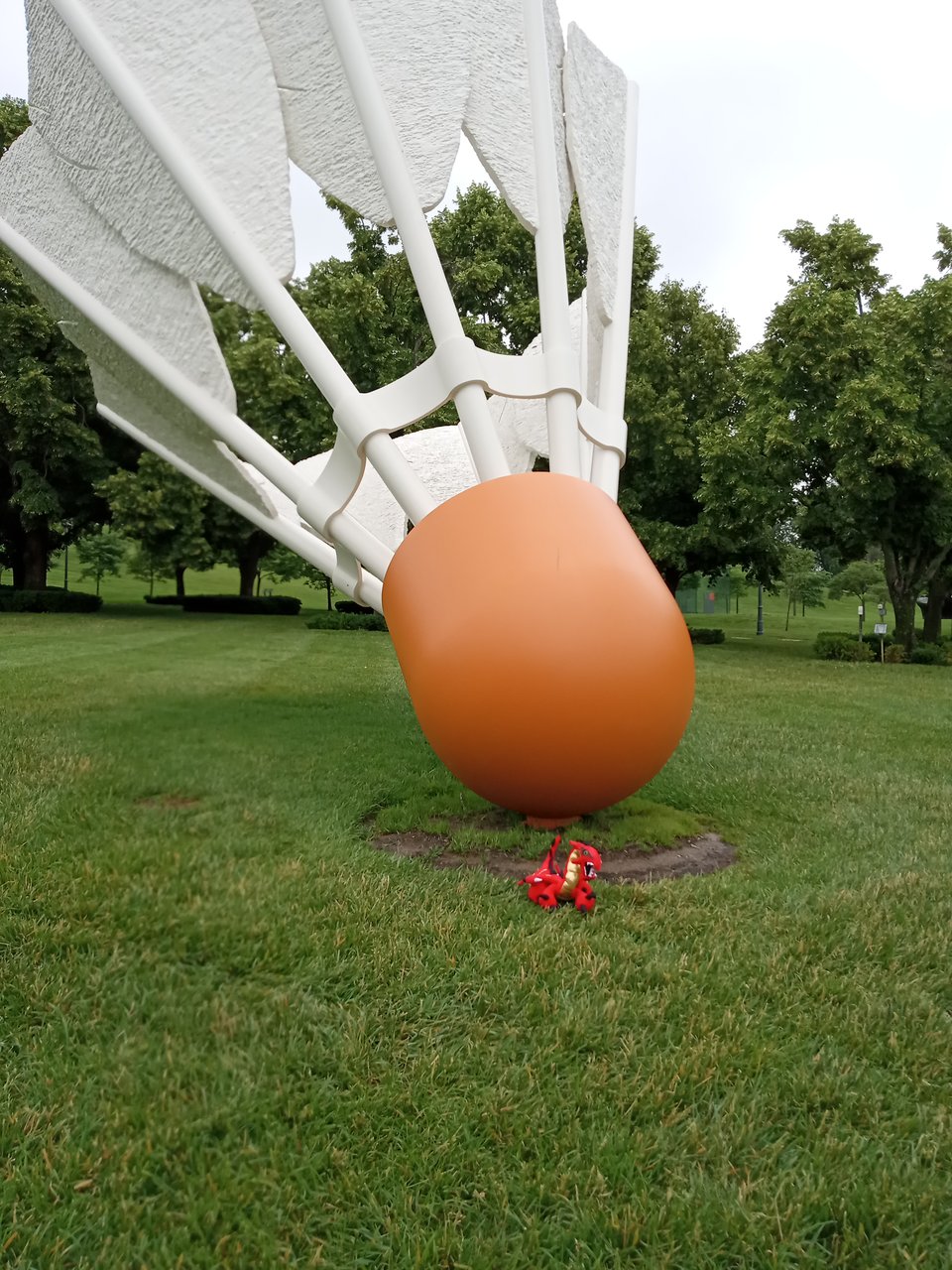 A very large shuttlecock with an orange base on a green lawn. Tiny red and gold dragon plushie for scale.