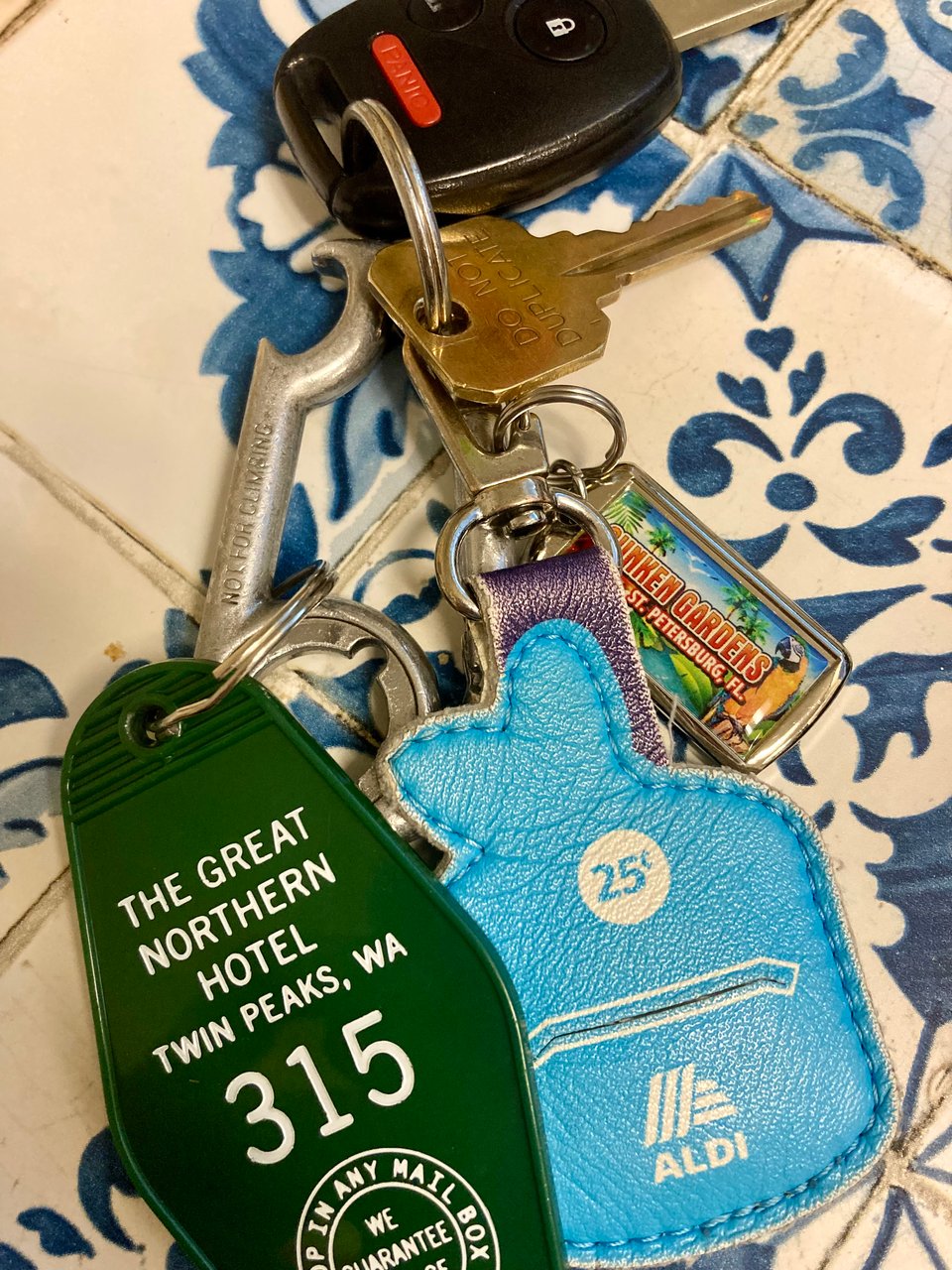 keys laying on a blue tiled table. besides a car and house key, there are three keychains: a green Great Northern Hotel room fob, a blue Aldi quarter-keeper, and a Sunken Gardens license plate.
