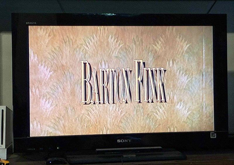 A photo of my television, which in this instant displays the title of the movie Barton Fink, against a background of ugly wallpaper.