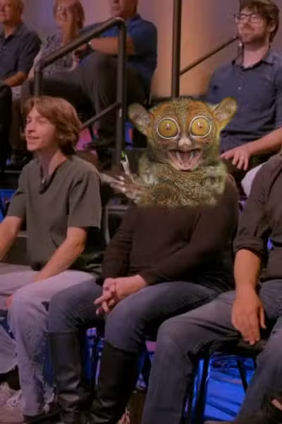 Three people sitting in an audience; the middle has a cutout of a tarsier face over their head