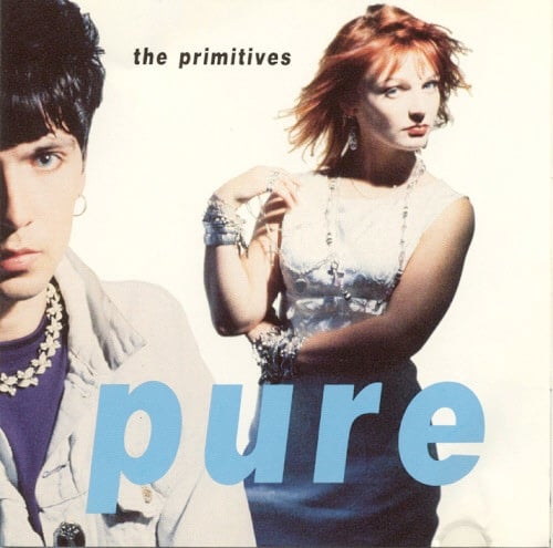 A color photograph of two members of The Primitives, both looking at the camera. One is a white man with short black hair, an earring, and necklace made of metal butterfly shapes. He is close to the camera and the left side of his face is cropped off. To the right and behind him is a white woman with red hair in a white dress. Her left arm is wrapped around her waist and her right arm is lifted so she's touching her right shoulder with her right hand. The background is white.