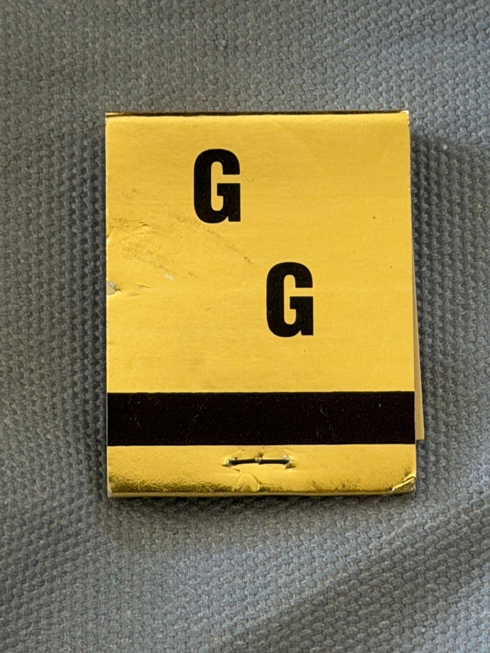 a gold matchbook cover that says G G in black font in a diagonal on a light blue canvas material