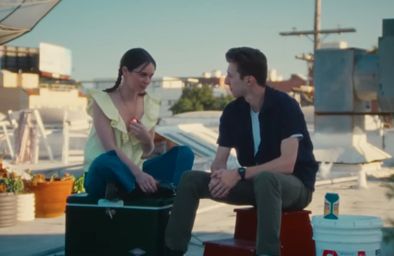 A young woman and young man sit on a rooftop on a sunny day. They are looking at each other. The woman is fingering a shiny pendant that she wears around her neck.