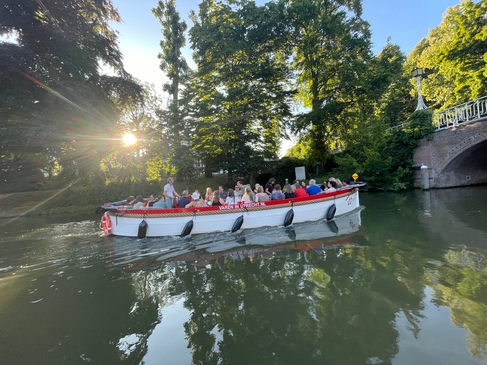 A river motor boat filled with people along the canals of Utrecht. The sun shines through the trees on the other side of the canal.