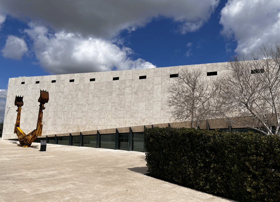 A light stone building against a blue sky with white clouds, with an art installation of the arms of two back hoes