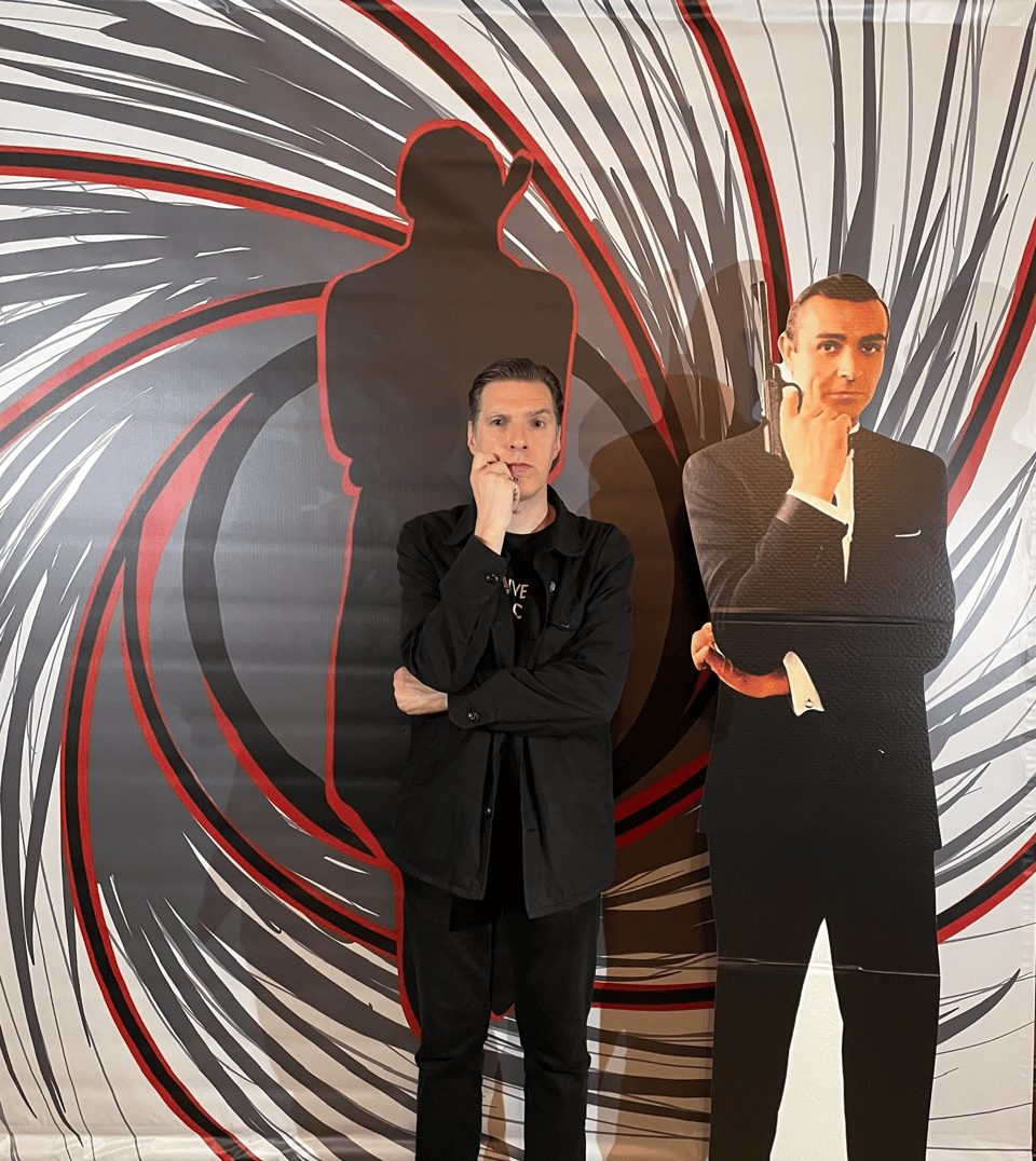 A man stands in front of a photo op next to a cardboard cut-out of Sean Connery as James Bond