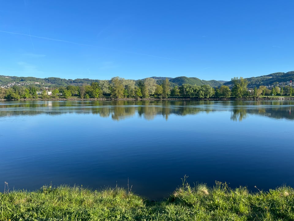 Looking across a wide river which has a mirror finish, reflecting the trees and blue sky on the other side.