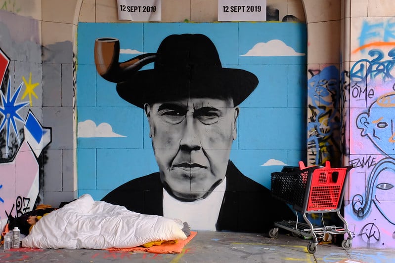 Image by Boudewijn on Flickr. "The graffiti on an outside wall is presumably an image of René Magritte. In front of it is the "living room" of two people. They are sleeping there. The shopping cart is there to transport whatever possessions they own."