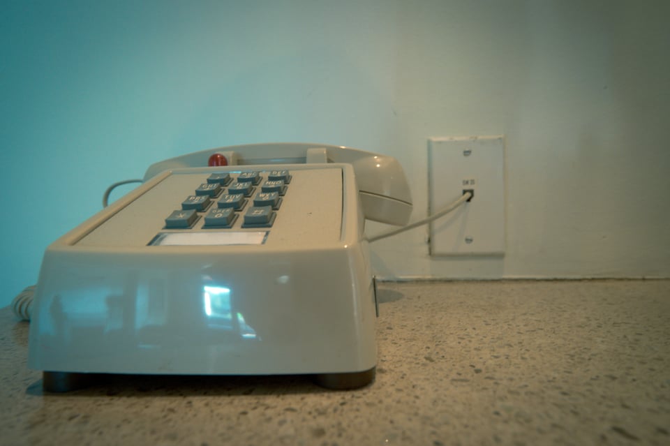 Old 1980s landline phone with 12 button keypad, biege, on a beige-brown countertop, plugged into a wall phone outlet