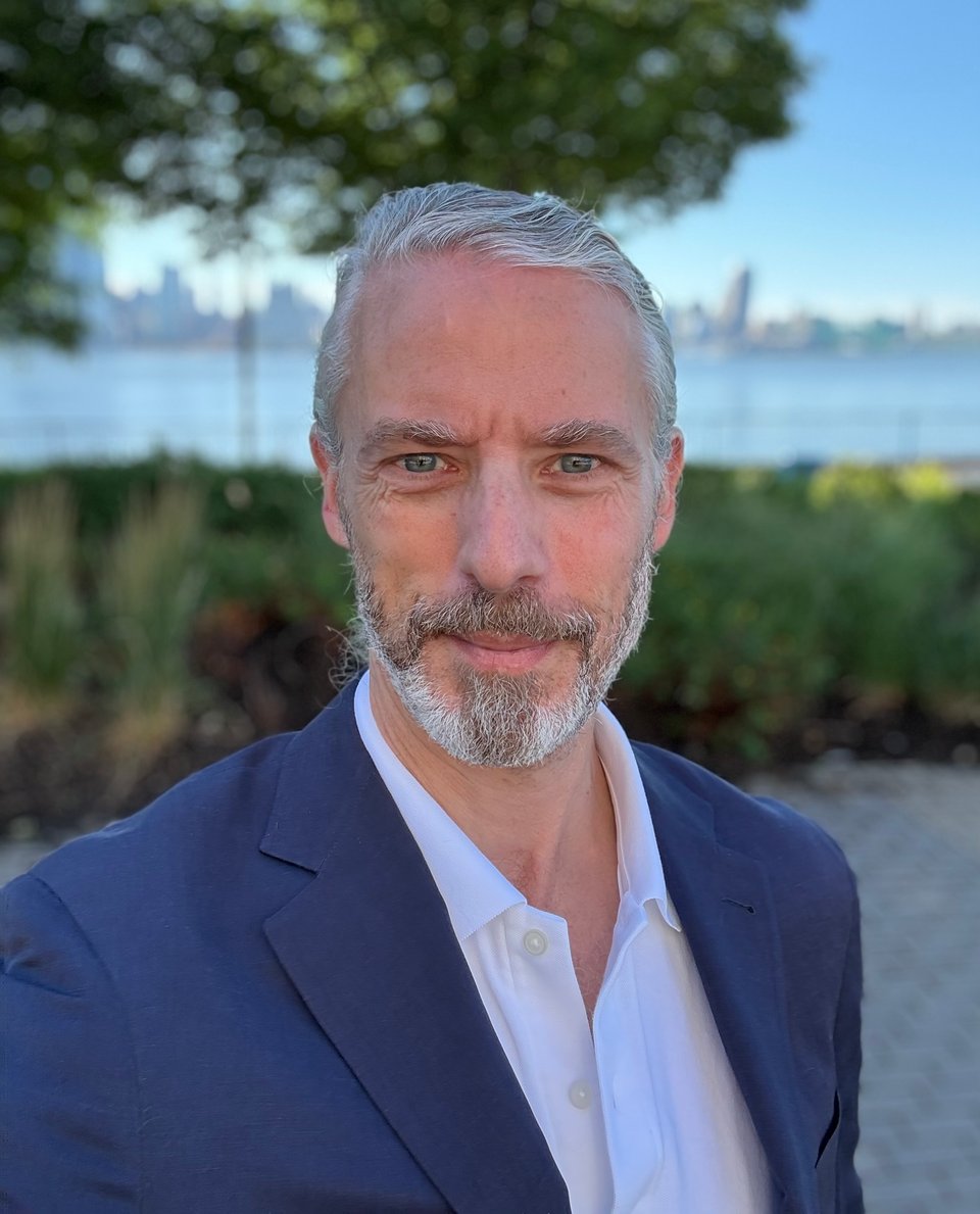 portrait-mode photo of white man in blue suit and white polo shirt smiling at camera with Hudson River and lower NYC skyline in background