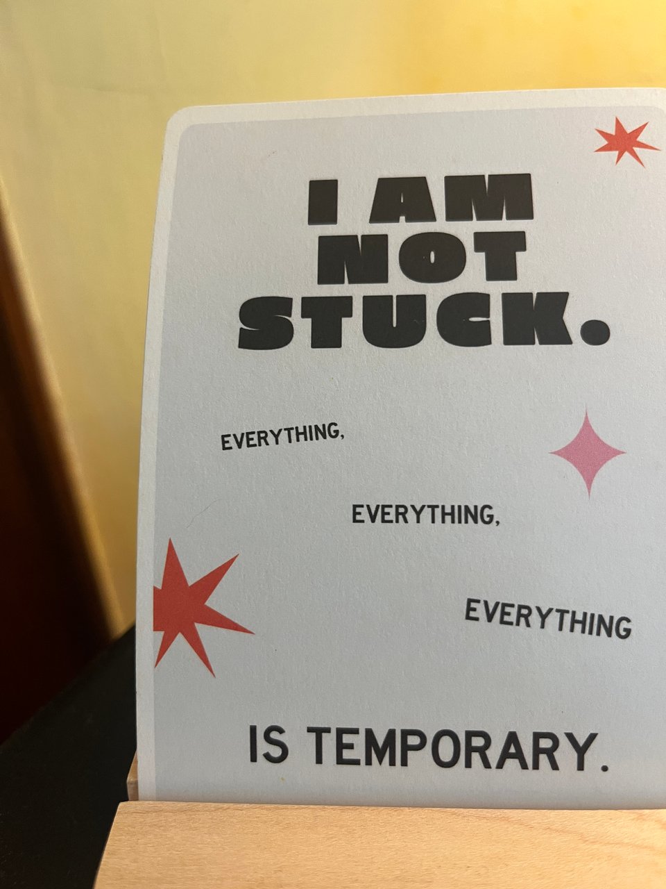 close up view of an astrological affirmation card (grey with black letters and pink and red starburst shapes) that reads, "I am not stuck. Everything, everything, everything is temporary."
