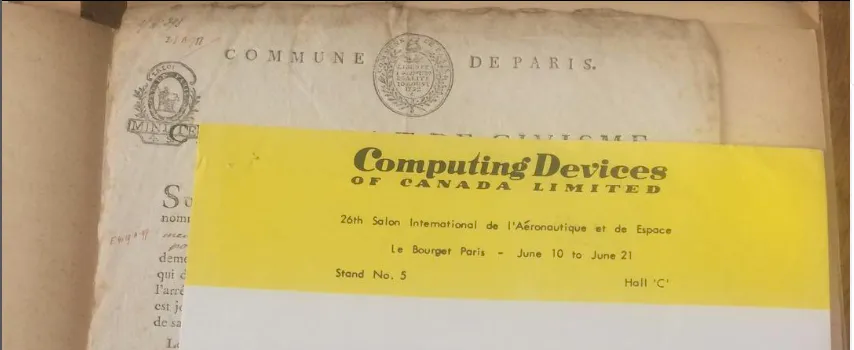 Photograph of two archival papers, once a bureaucratic document for the Commune de Paris, the other a yellow press release for Computing Devices of Canada Limited