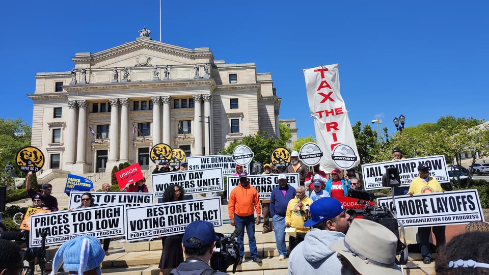 Protest signs provide a background to the speaker at the May Day protest in Newark, NJ. The Old Essex County Courthouse is in the background, with the deep blue sky composing the top of the image.
The signs cover a variety of topics: No more layoffs. Restore Voting Rights Act. Tax the RIch. Prices too high, Wages too low.