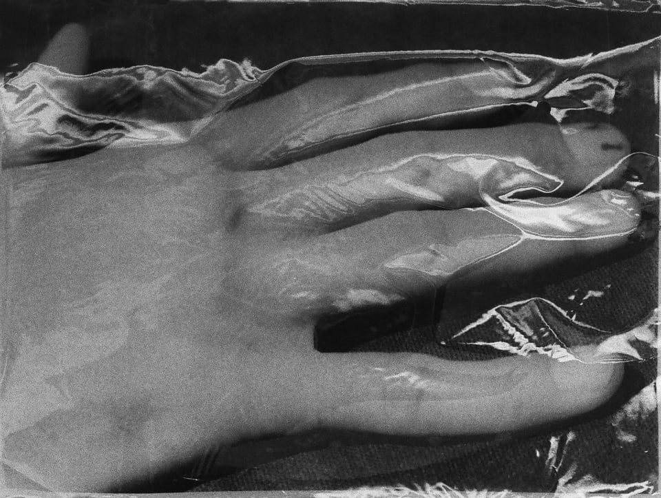 A black-and-white photograph of a hand pressed against a sheet of plastic or cellophane, the material catching light and creating a crumpled, translucent surface around the fingers and palm.