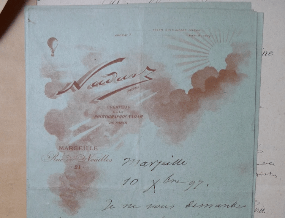 A light blue piece of stationary with a letterhead design printed in red. The image shows clouds with a sun and balloon, on top of the image is the name Nadar and the address of the studio in Marseille.