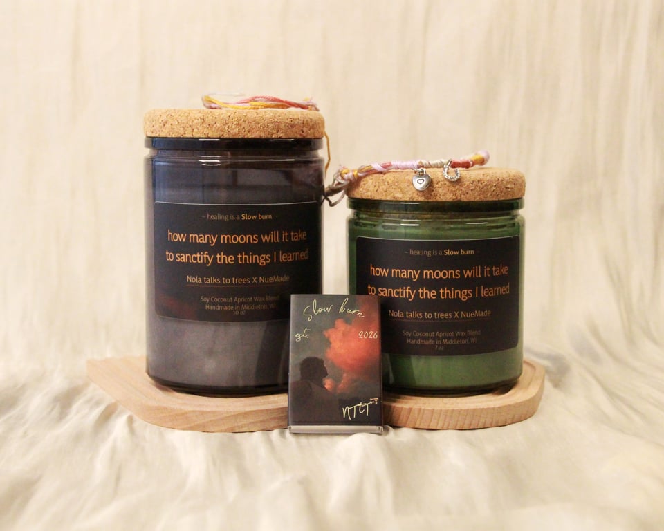 Two candles, one taller than the other, against a cream cloth background. A Slow burn matchbox is leaning against the candles and a friendship bracelet with charms rests on top of the candles. Both have a label with the lyric "how many moons will it take to sanctify the things I learned."