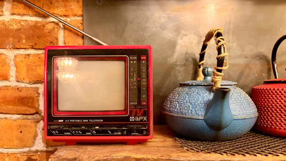 On a shelf in front of a red-brick wall: a red-framed 4.5-inch TV set with a retractable antenna, next to a blue and a red cast-iron teapot