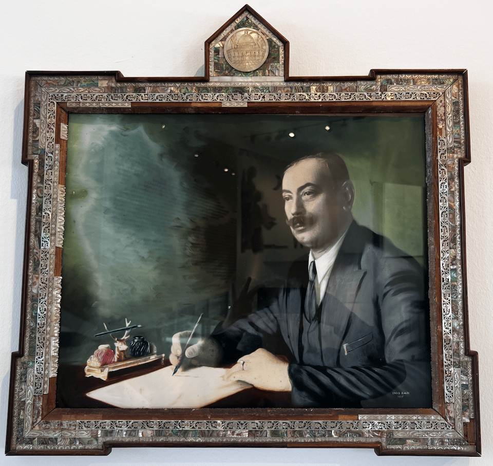 A portrait of a man sitting at a desk, holding a pen near a piece of paper on the desk, within an intricate mother-of-pearl frame