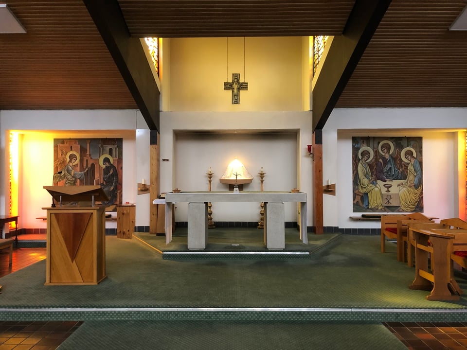 The inside of the church. A simple stone alter sits on a green dais that is set between two large tapestries. A large sloping ceiling, clad in wood, dips down towards the alter. To either side of the tapestries, amber light is flooding in. Above the alter, the roof rises, and two more side windows illuminate the cross on the wall. Even the lectern looks midcentury.