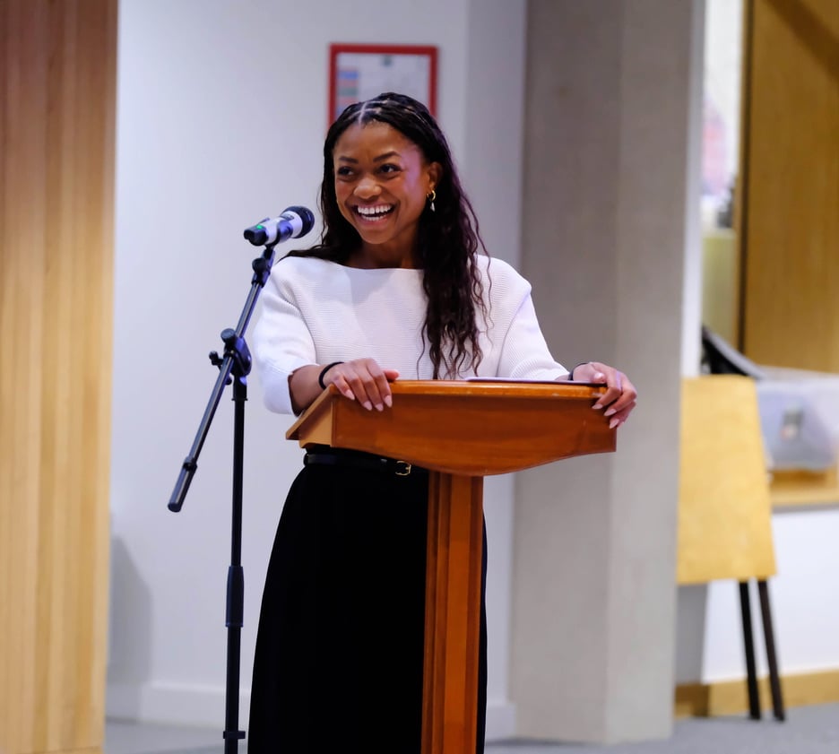 Nadia laughs broadly while standing behind a wooden lectern, resting both hands on its edges. A microphone stands beside her, and she wears a white top with black wide-leg trousers.