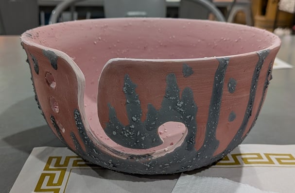 A yarn bowl that has been painted with a reddish glaze on the inside and outside, with streaks of a greyish glaze with irregular flecks throughout on the exterior