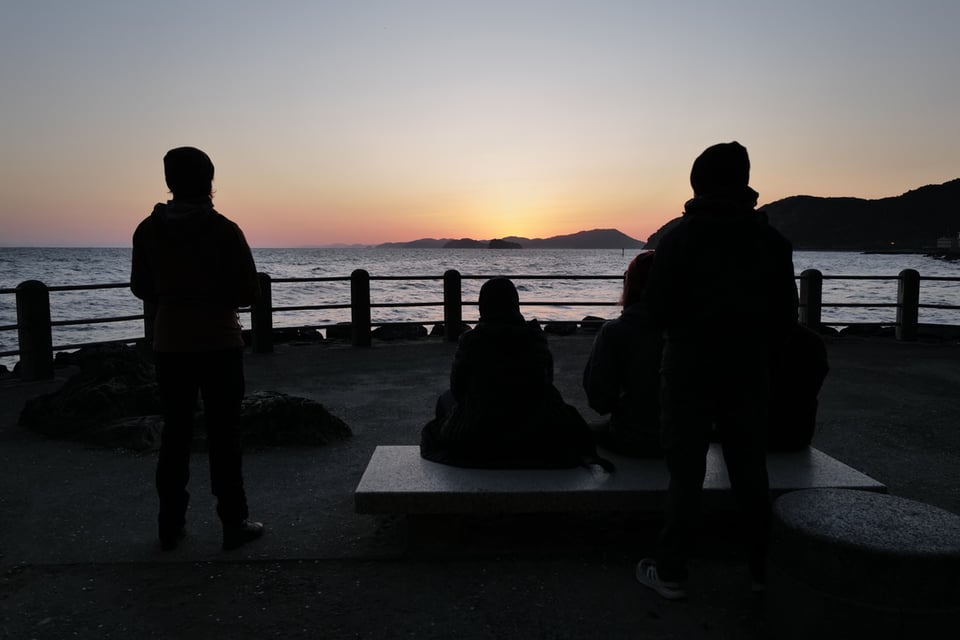 Four silhouetted figures on a seaside promenade watching the sun rise behind island outlines on the horizon, the sky layering from deep orange at the waterline through pink to pale grey above