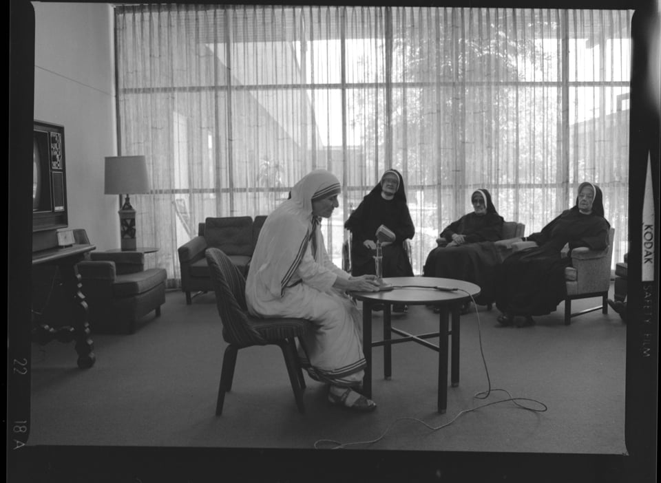 An elderly woman in white sari sitting in front of an old-fashioned microphone; three more women in black habits sitting in background.