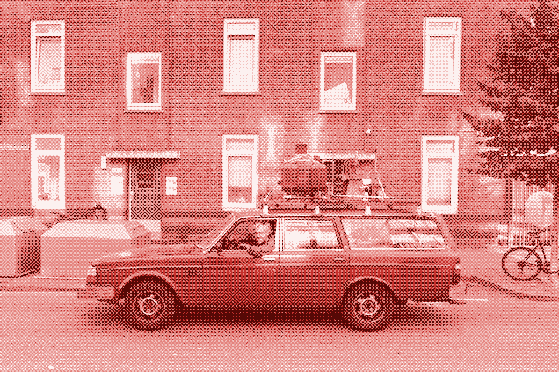 A picture of a Volvo station wagon, looking fairly old, perhaps in the 80s. It is parked at the side of the road and pointed to our left. In the driver seat, there is a smiling man who is leaning out the window looking at us. On top of the car is a large contraption, about 4 feet tall and 6 feet long, that looks like three large cam canisters strapped together with a sort of stove pipe on top. In the background is a row of brick apartment houses. The picture is tinted a light rust color.
