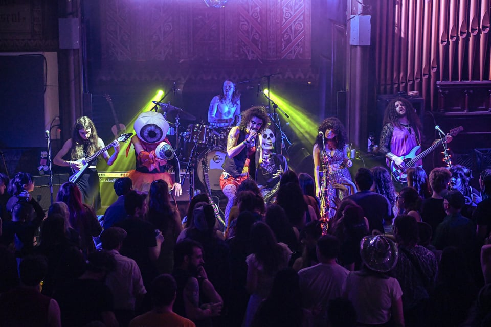 Six colorfully attired musicians perform onstage, with pipes from a large organ at the back right and yellow lights shining from either side of a drum riser.