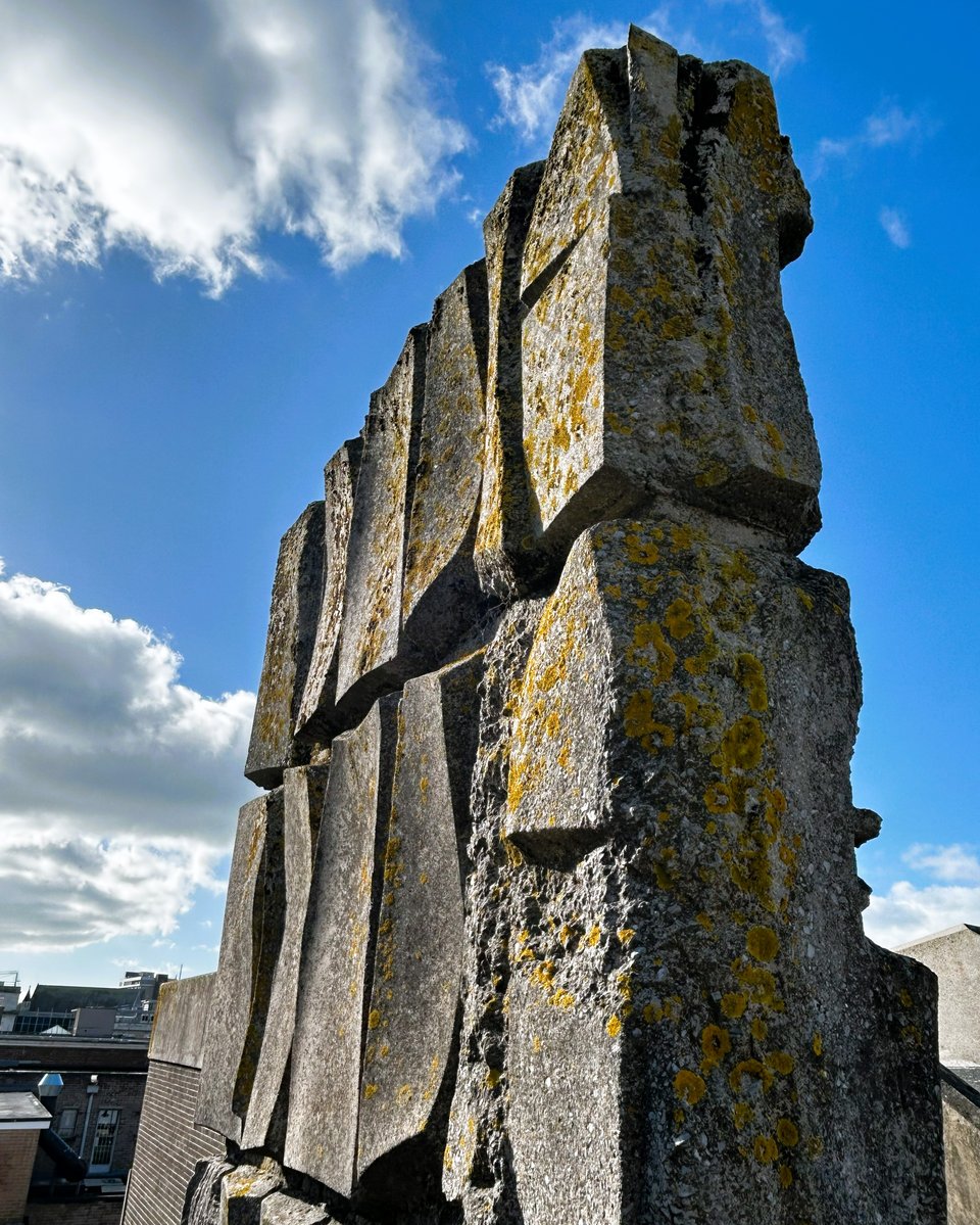 Two cast concrete blocks forming the central column of a staircase as it emerges on the top floor of a carpark. The concrete has been shaped to look like folded rock. It's smooth, apart from at one end where the concrete seems to be crumbling. It's a dark grey, like granite, with orange blooms of lichen all over it. There's a blue sky with high white clouds behind it.