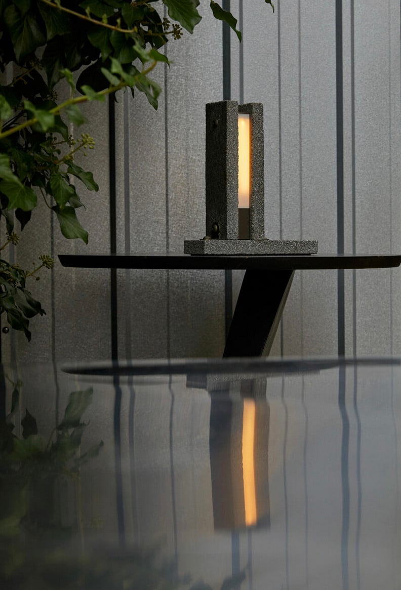 A picture of the lamp. It is a tall rectangular shape; on the left and right side are long strips of concrete, and in the middle is a softly glowing yellow light. The lamp is mounted on a wall and reflected in a pool of water beneath it
