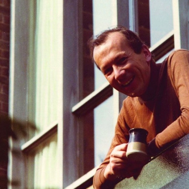A middle-aged white man wearing a brown poloneck sweater and leaning out of a modern window. He's holding a 1960s or 1970s stoneware coffee cup