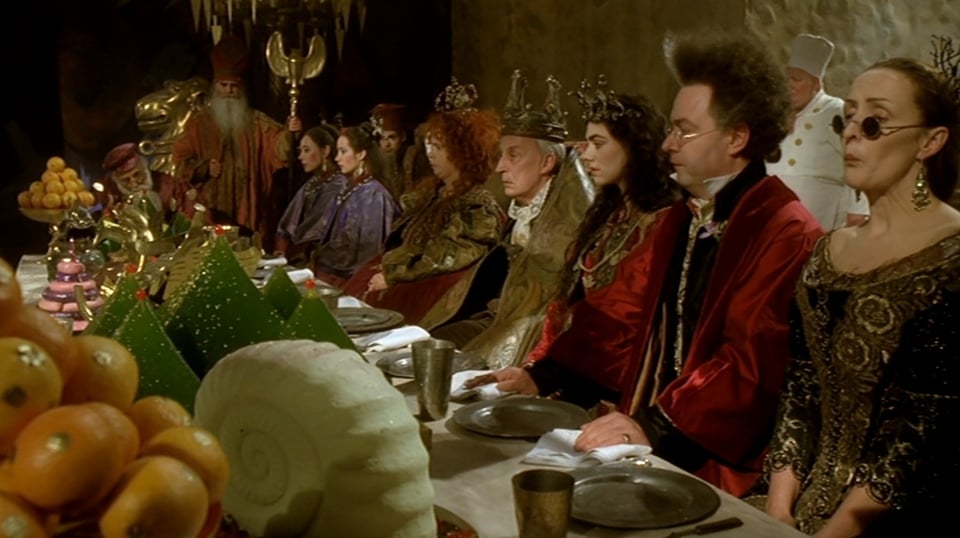 A row of outlandishly dressed nobles sitting a long banquet table, which is laden with unlikely-looking foods. They are all staring into space, as if consumed in thought.