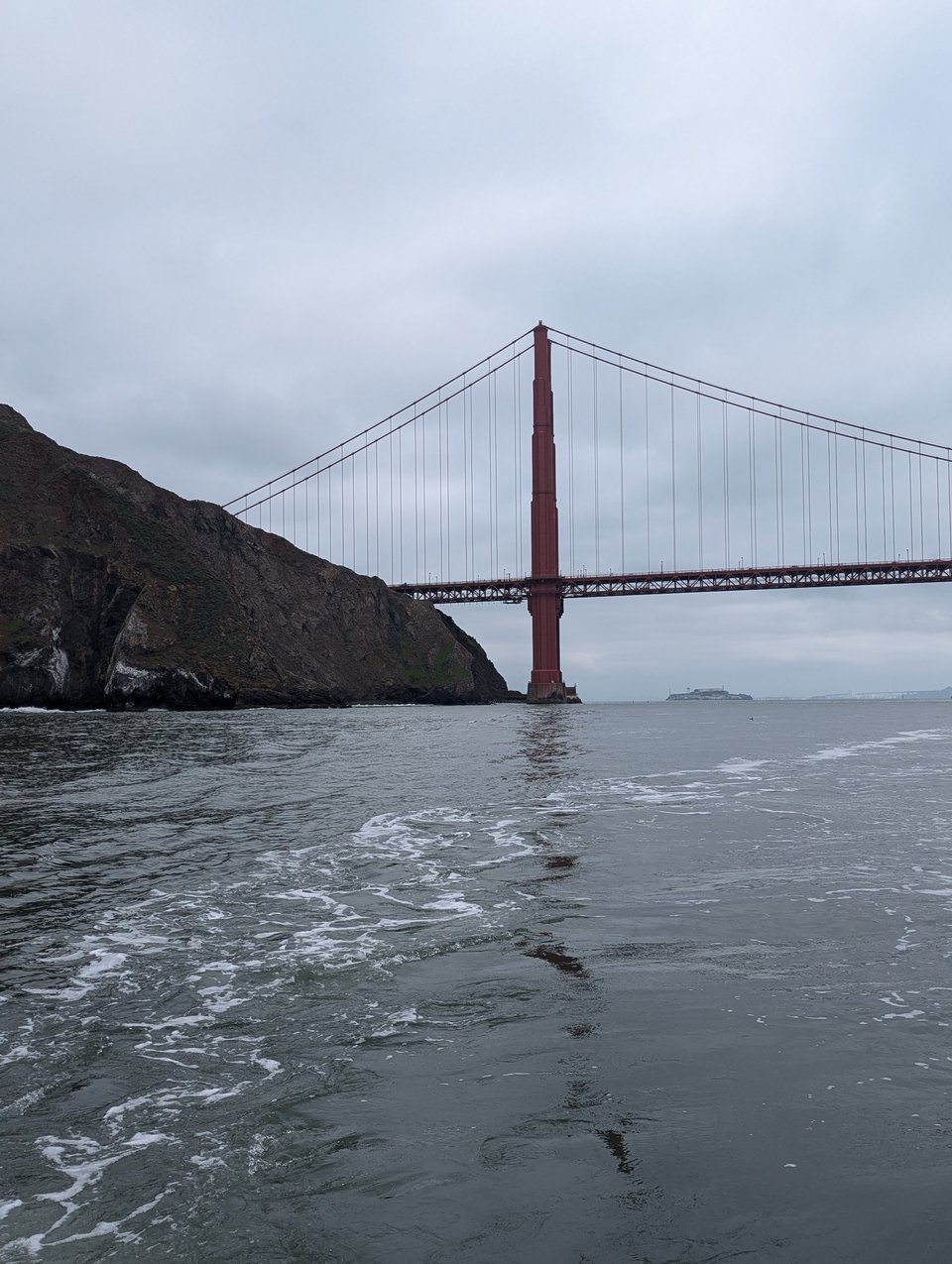 The grey ocean along grey land under a grey sky. The Golden Gate Bridge stands overhead. Its jagged reflection shows in the water.