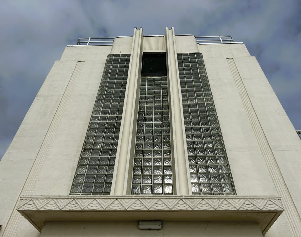 A cream colour staircase block. It has three full length windows made of glass blocks: the central one has a panel with an etched Deco drawing of a diver at the top. The concrete porch has decorative details.